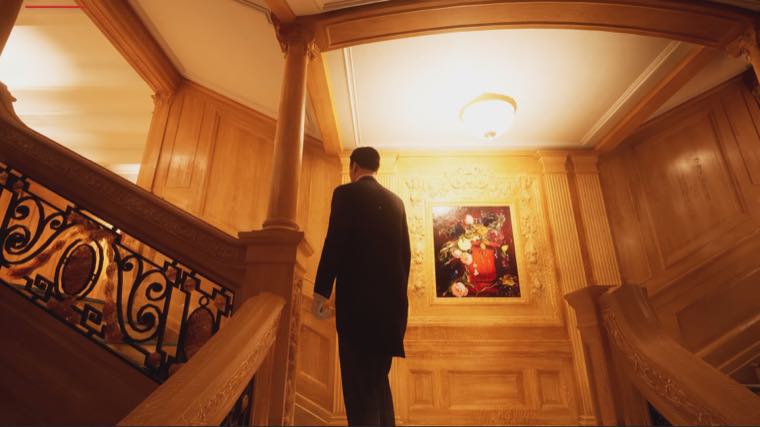 The oak-panelled Grand Staircase, a man in a dark suit standing on the landing beside a framed still-life painting